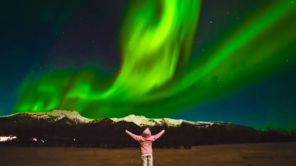 Dreamy girl in pink jacket watch the northern lights show alone in meadow field. Romantic Woman on Icelandic spring night landscape. Iceland travel chasing northern lights concept
