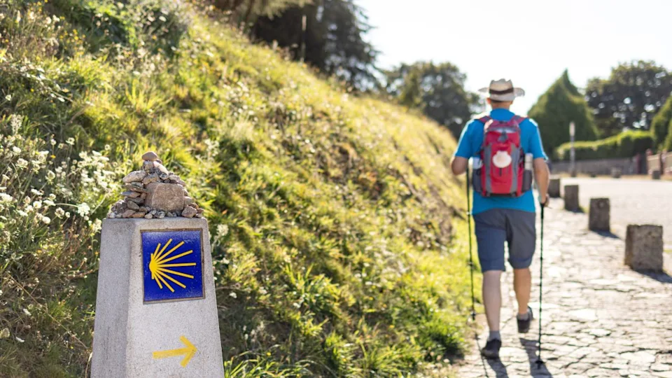 Pilgrim walking to Santiago de Compostela in Camino de Santiago pathway guided by milestone sign with scallop shell and arrow indicating the way