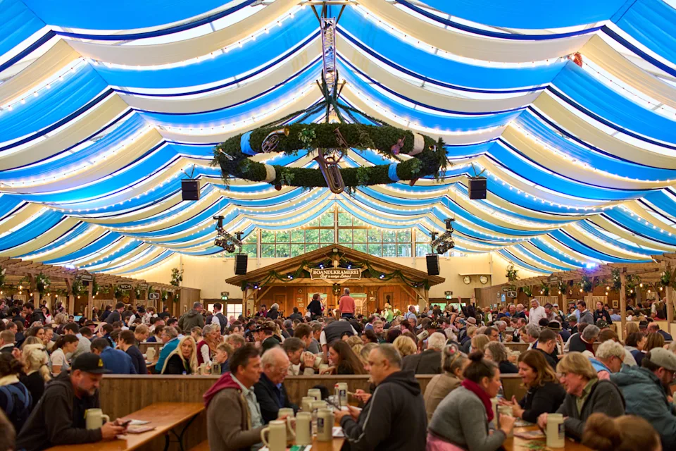 Large crowd enjoying food and drinks under a festive tent with striped, draped ceiling at an Oktoberfest-style event
