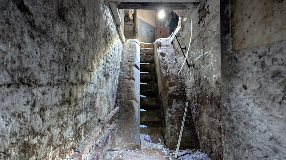 Stairway descent into dark home basement. Eerie ancient staircase like steps into tomb ruins, crypt or catacombs. Stone cut crumbling brickwork walls. Inside spooky abandoned cellar or archaeology dig site remains. Shadows cast from light in darkness. Derelict dungeon setting underground is gloomy hallway leading up to stairs. Mysterious lost brickwork passageway feels ghostly and haunted hidden beneath house. Old empty tunnel