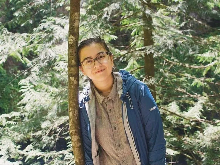 The author leans against a thin tree with more trees behind her