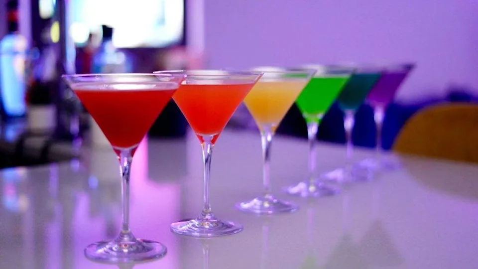 Colorful cocktails in martini glasses arranged in a rainbow gradient on a glossy surface.