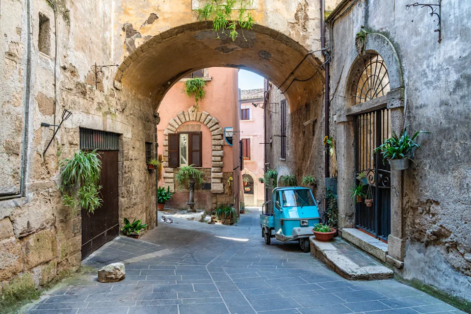 Charming narrow street in an ancient town, featuring stone buildings, a small three-wheeled vehicle, and lush potted plants