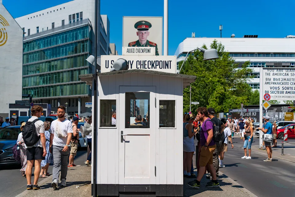 People gather around a historic U.S. Army checkpoint building in a busy urban area, likely a popular tourist attraction