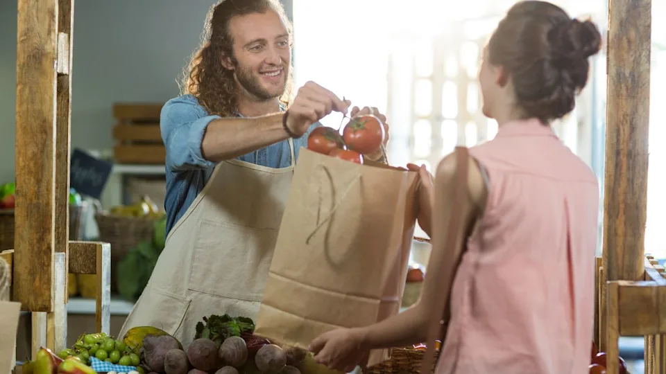 Smiling vendor handing a bag of vegetables to woman at grocery store