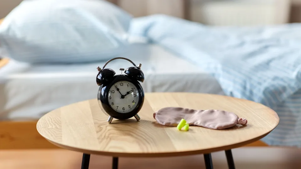bedtime and morning concept - close up of alarm clock, earplugs and eye sleeping mask on night table at home