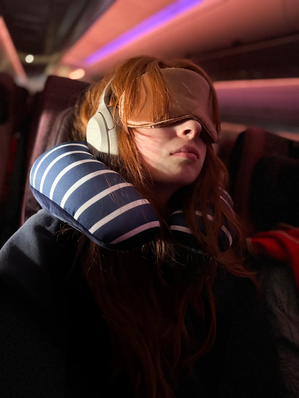 Stock photo showing close-up view of a red haired female traveller sat in airplane cabin sleeping, whilst wearing an eye mask, wireless, noise cancelling headphones and leaning against a neck pillow, in a plane window seat. The airplane cabin has been illuminated by pink neon, mood lighting designed to soothe boarding passengers.