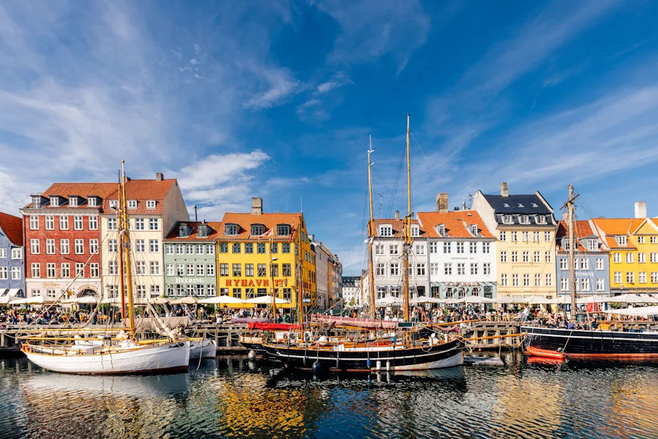 Boats docked in a canal with colorful historic buildings lining the water in a bustling neighborhood, likely Nyhavn, Copenhagen