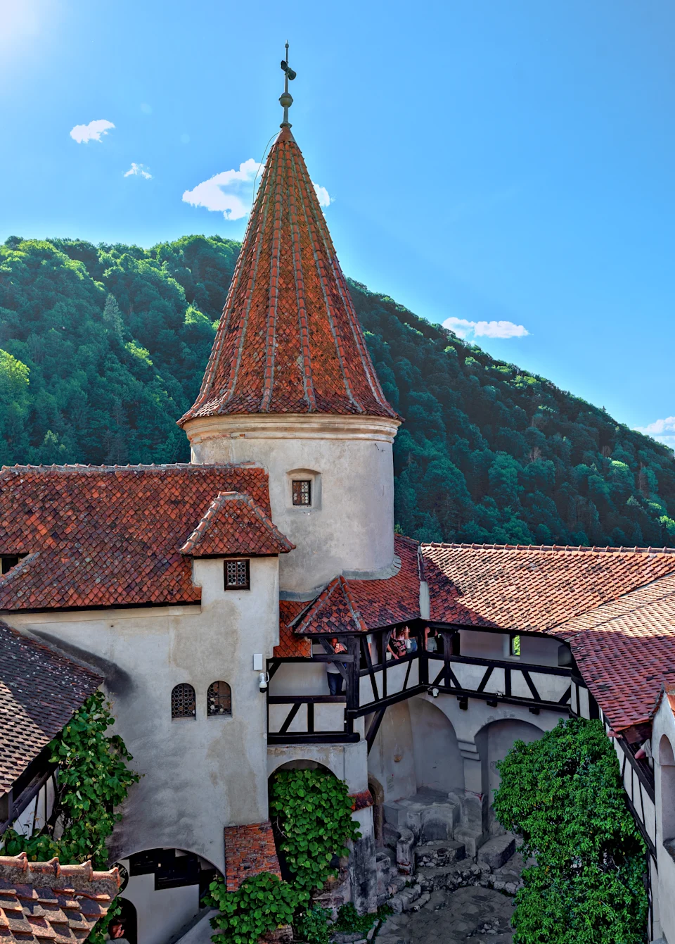 Castle courtyard with a tall, pointed tower and scenic mountain backdrop. Visitors are exploring the historic architecture