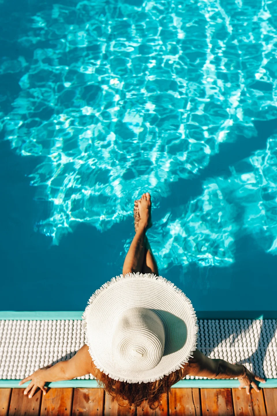 Person relaxing by a pool, wearing a wide-brimmed hat, legs dipped in water. Top-down view suggests leisure and work-life balance