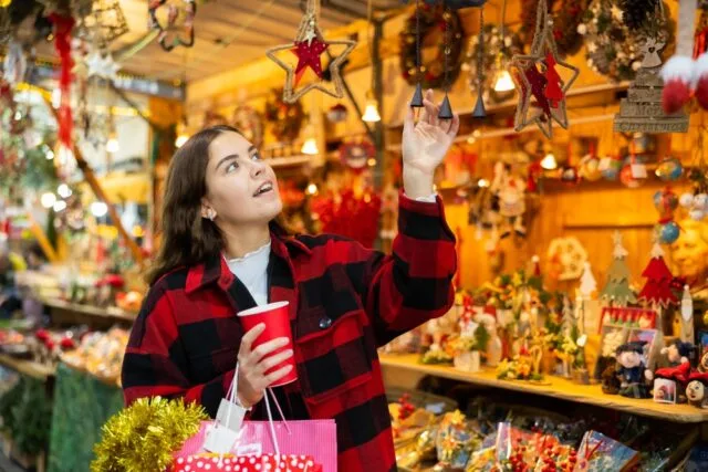 Young woman walking at colorful Christmas Street market with paper cup of warm tea in hands, looking for holiday decorations.