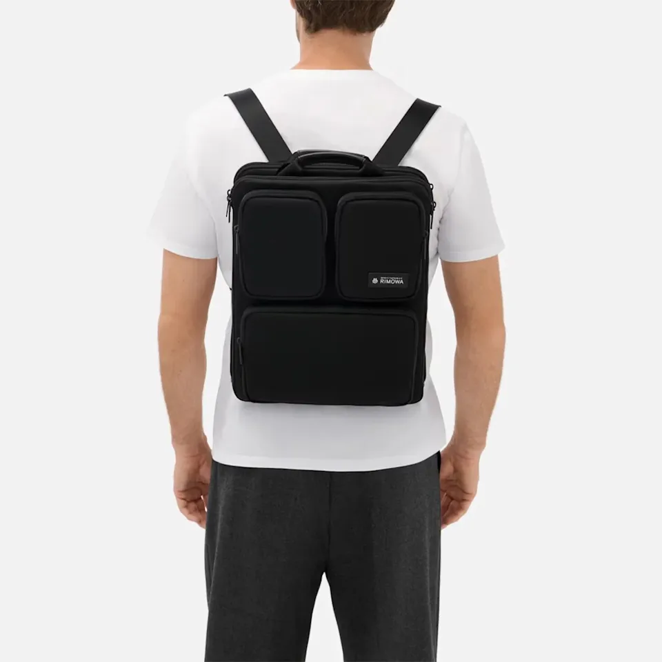 Black rectangular backpack with multiple zippered compartments worn by a person in a white shirt and dark pants.