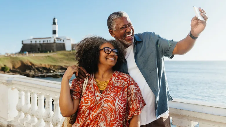 Two seniors smiling taking a selfie in front of a lighthouse near the water.