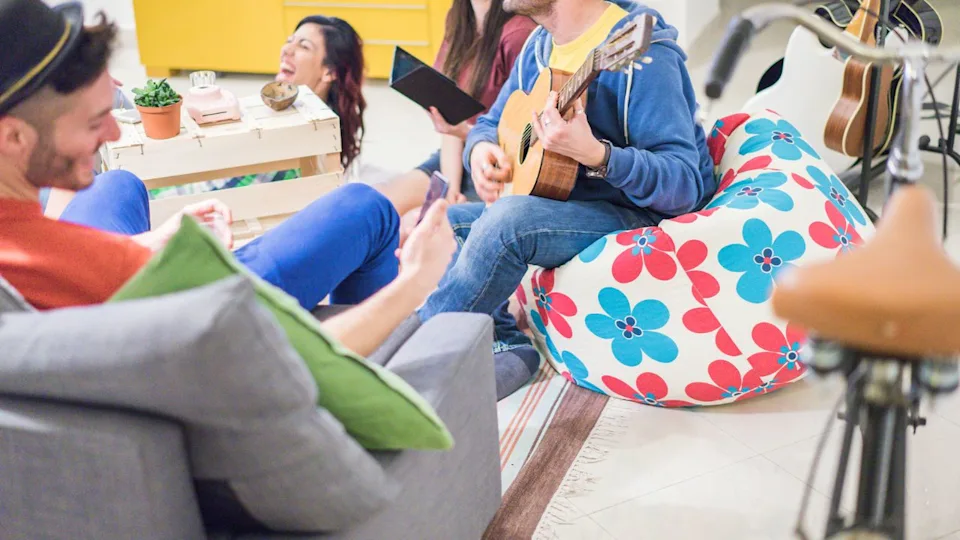 Group of trendy friends having fun in hostel living room - Happy young people enjoying time playing music and watching videos on smartphone - Focus on man left hand guitar - Warm contrast filter