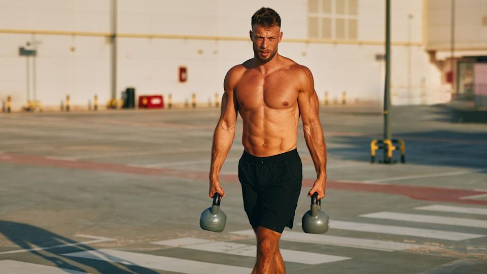Muscular male walking with kettlebells outdoors.