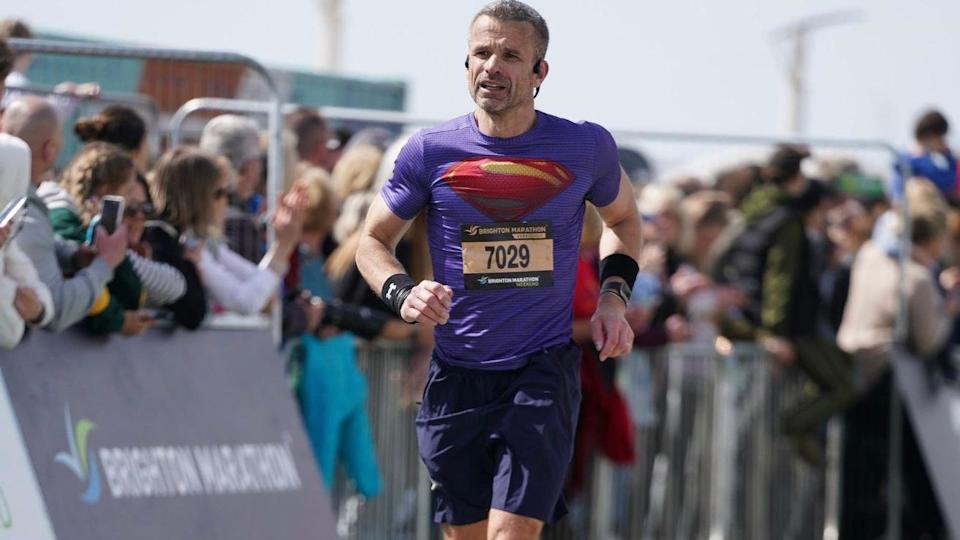 A man wearing a purple sports t-shirt and navy shorts. He is wearing bright blue trainers. He is running at a race with lots of spectators at the side.