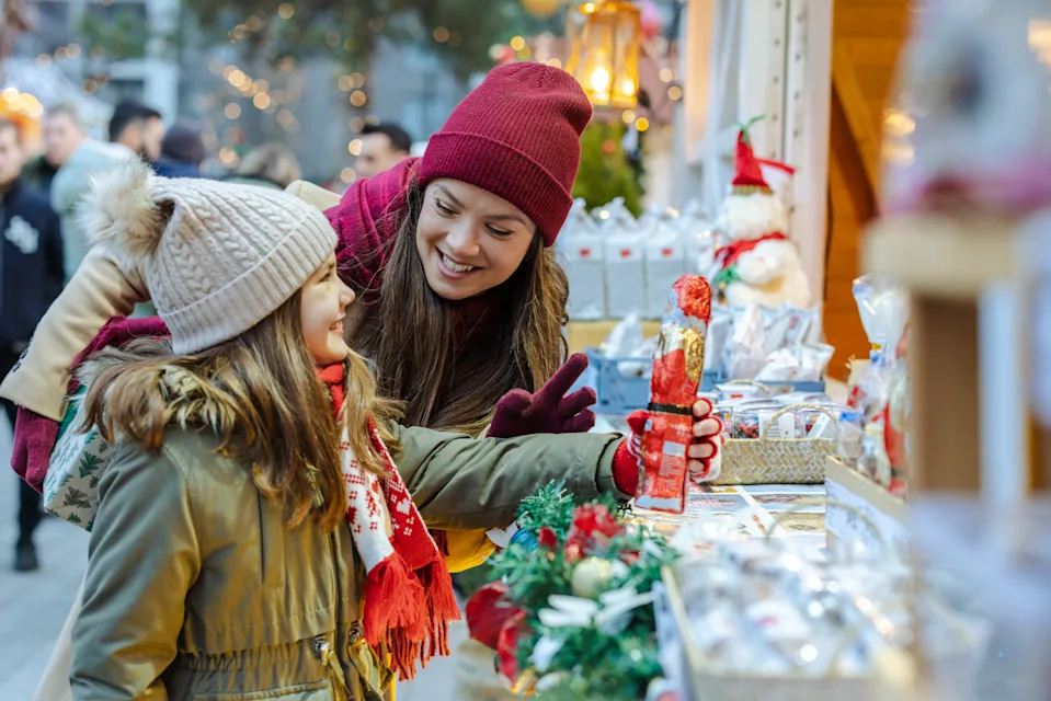 A mother helps her daughter pick a festive treat at a bright Christmas market stall. The holiday lights glow as they smile, share warmth, and create cherished family memories.