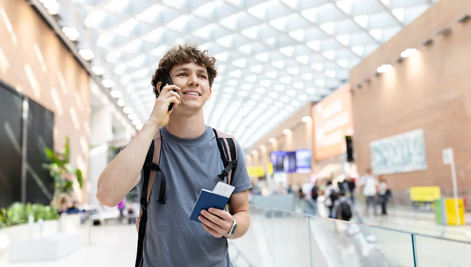 Young Caucasian man with backpack holding passport and phone while talking at airport. Represents boarding routine, youth travel lifestyle and positive moments