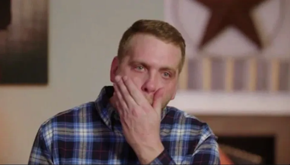 Person in plaid shirt appears emotional, holding hand to mouth in an interior setting with star decor on the wall