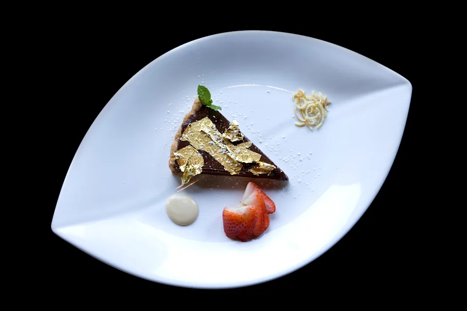 Slice of chocolate cake with gold leaf garnish on a white plate, accompanied by a strawberry and a dollop of cream
