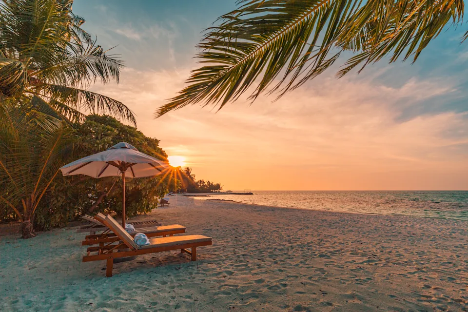 Beach scene at sunset with palm trees, sun loungers under an umbrella, and calm ocean waves reflecting the setting sun