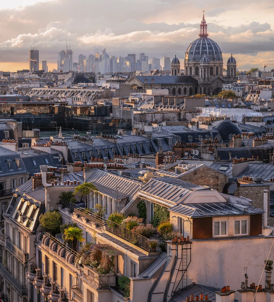 A scenic view of Paris rooftops with a historic dome in the background, framed by distant city skyscrapers under a cloudy sky