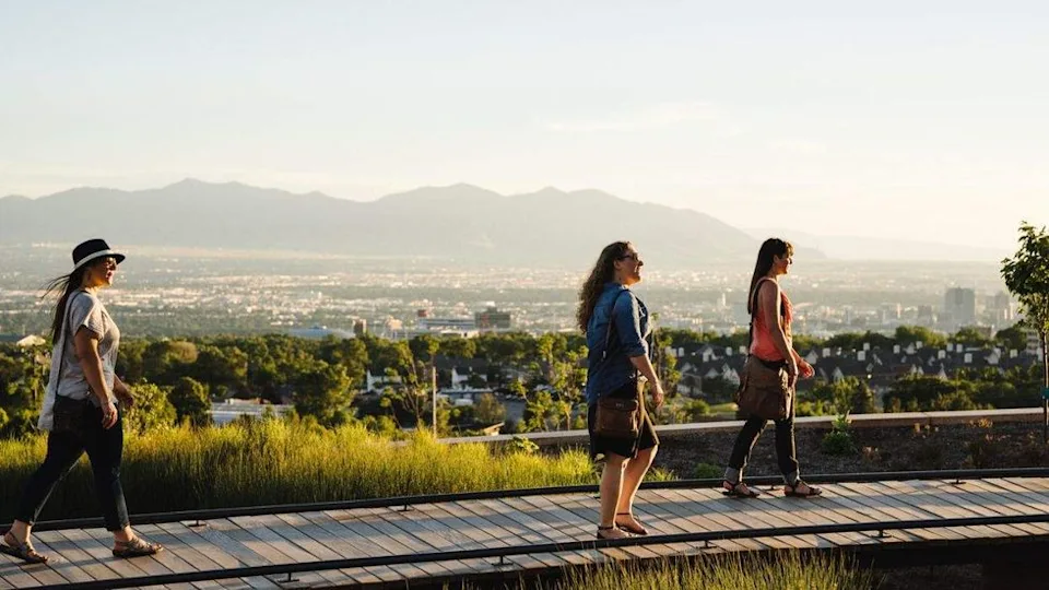 Three women walking on a boardwalk with city and mountains in the background.