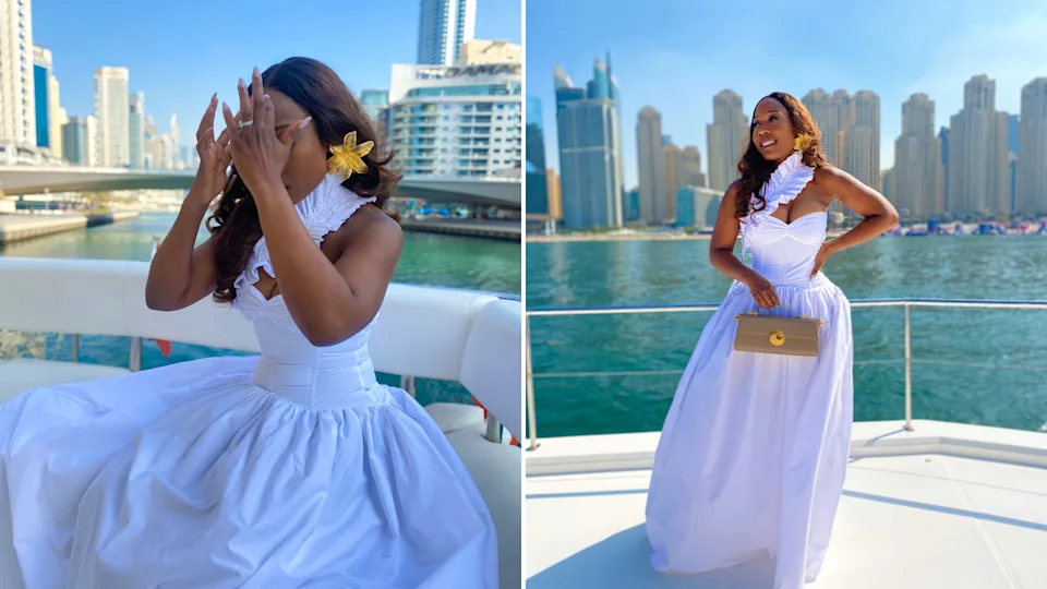 I enjoyed a yacht ride in Dubai Marina while wearing a white dress and holding a beige handbag with the city skyline behind me.