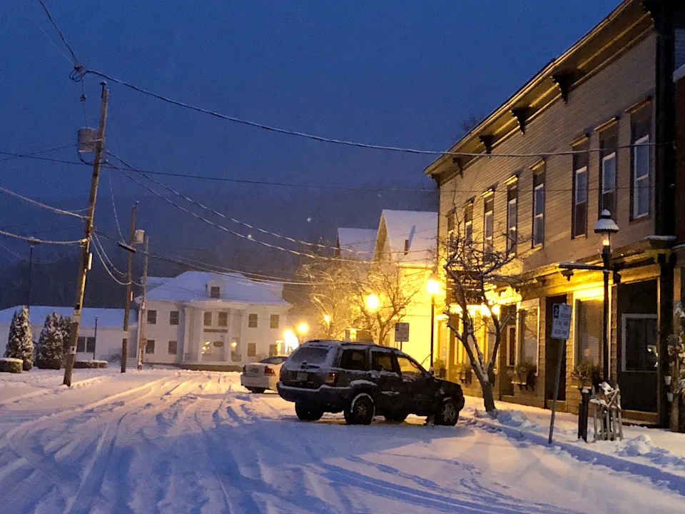 Snow-covered street in a quiet town, with a parked SUV and warm glowing streetlights illuminating the scene