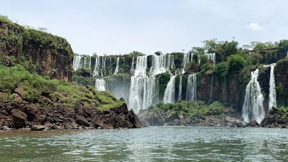 Iguazu Falls scenic view with powerful waterfalls, lush greenery, mist, and flowing river under cloudy sky. Iguazu National Park, Brazil Argentina border.