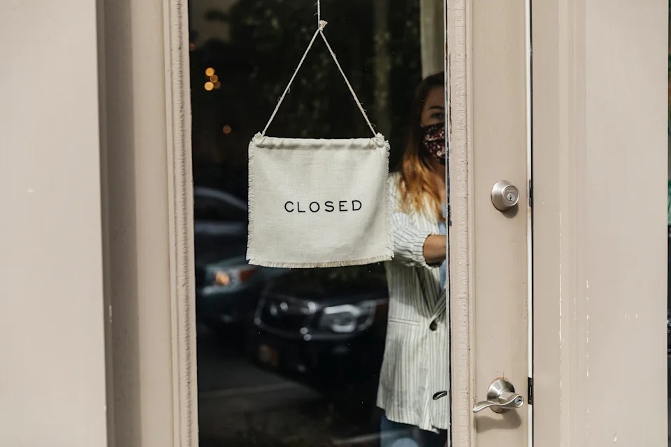 A person wearing a mask closes a glass door with a "CLOSED" sign hung on it, reflecting urban surroundings