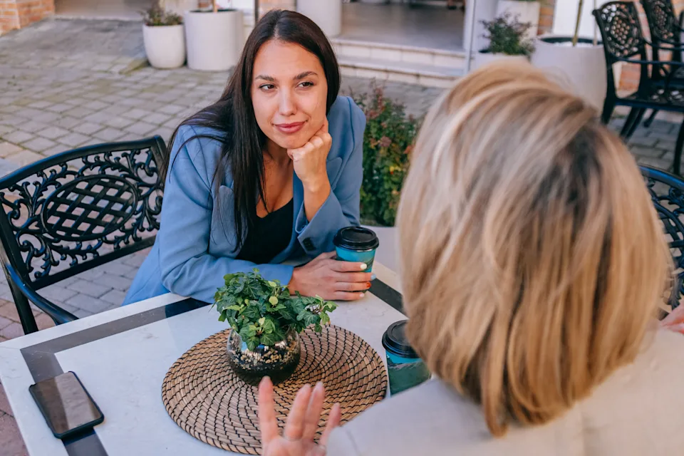Two people sit at an outdoor café table talking, each holding a coffee cup, with a plant centerpiece in a woven basket