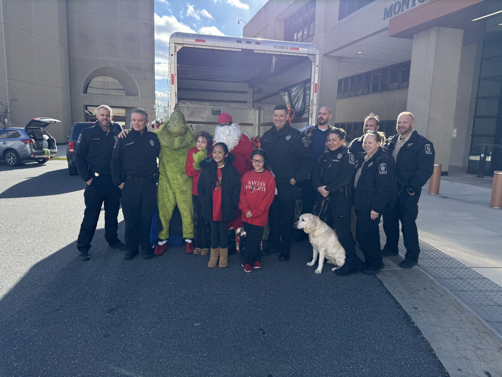 kids, police officers and people dressed as santa and grinch pose for group photo in front of box truck carrying toys for toy drive