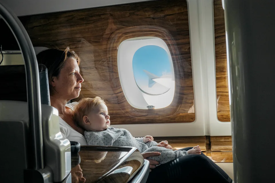 A mature woman sits in a business class seat on a flight with her baby daughter on her lap. They watch the TV (off screen) together.