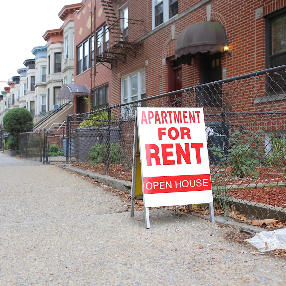 Sidewalk view of a residential street with a prominent "Apartment for Rent, Open House" sign near a row of brick buildings
