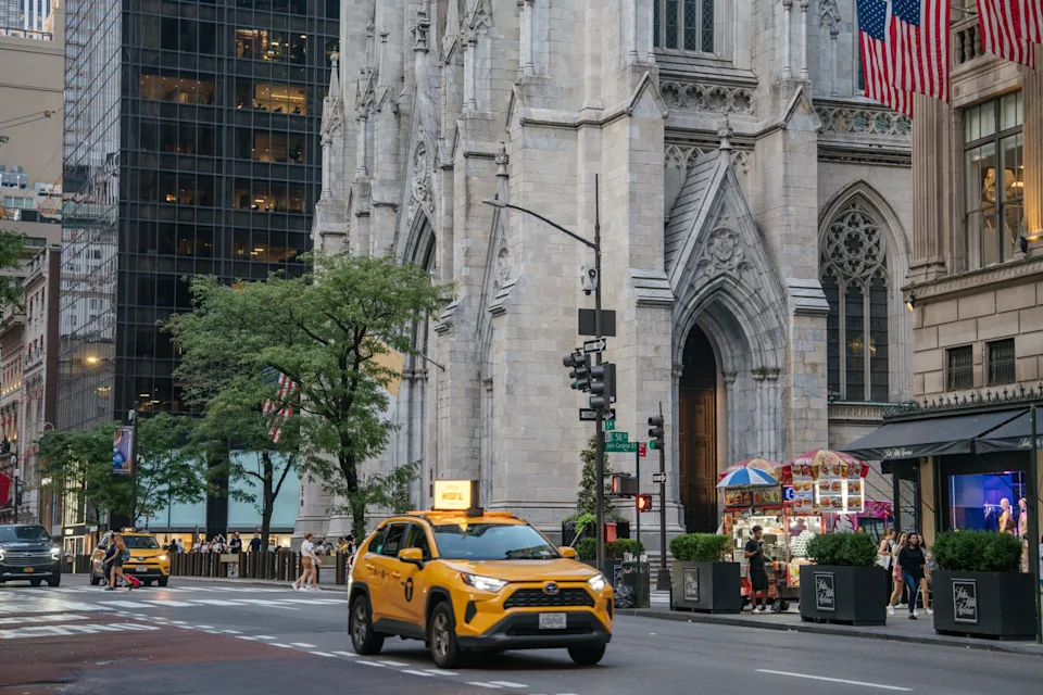New York City, New York, USA - 07.29.2024: A yellow NYC taxi drives past the grand entrance of St. Patrick's Cathedral on Fifth Avenue. The Gothic-style cathedral stands proudly beside modern