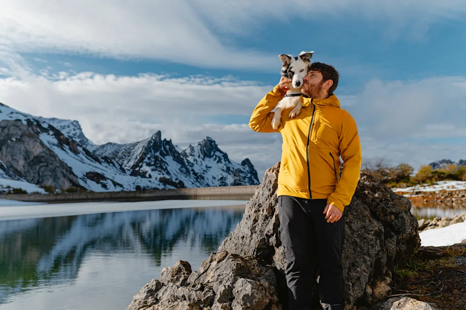 Happy man wearing a yellow jacket holding and kissing his dog on his shoulder, enjoying a winter hike