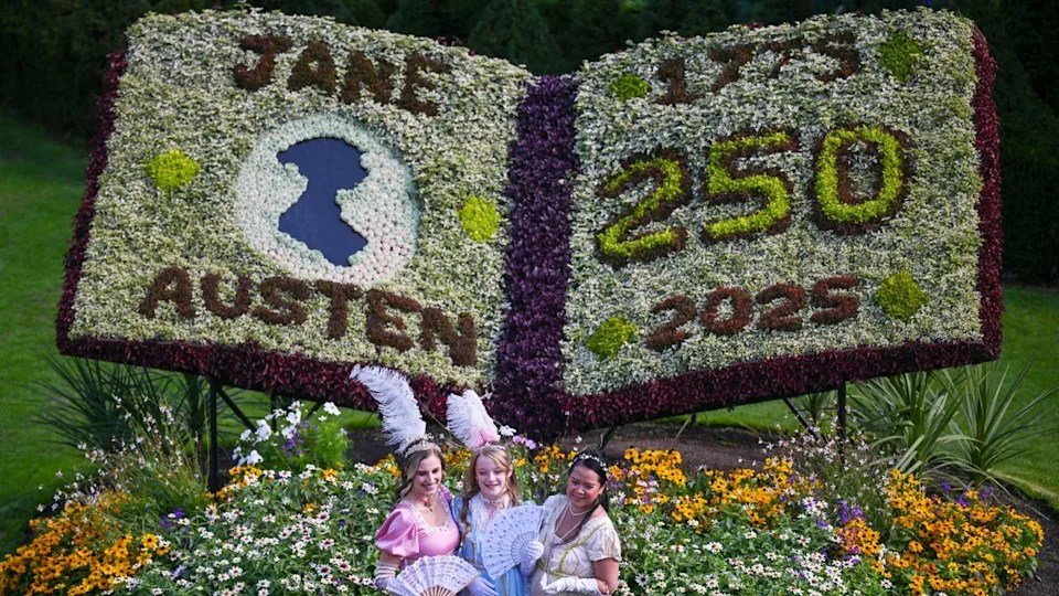 Three women dressed in Regency outfits stand in front of a floral decoration honoring Jane Austen.