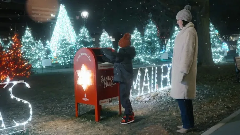 A mother watches her child post a letter to Santa surrounded by Christmas trees and lights