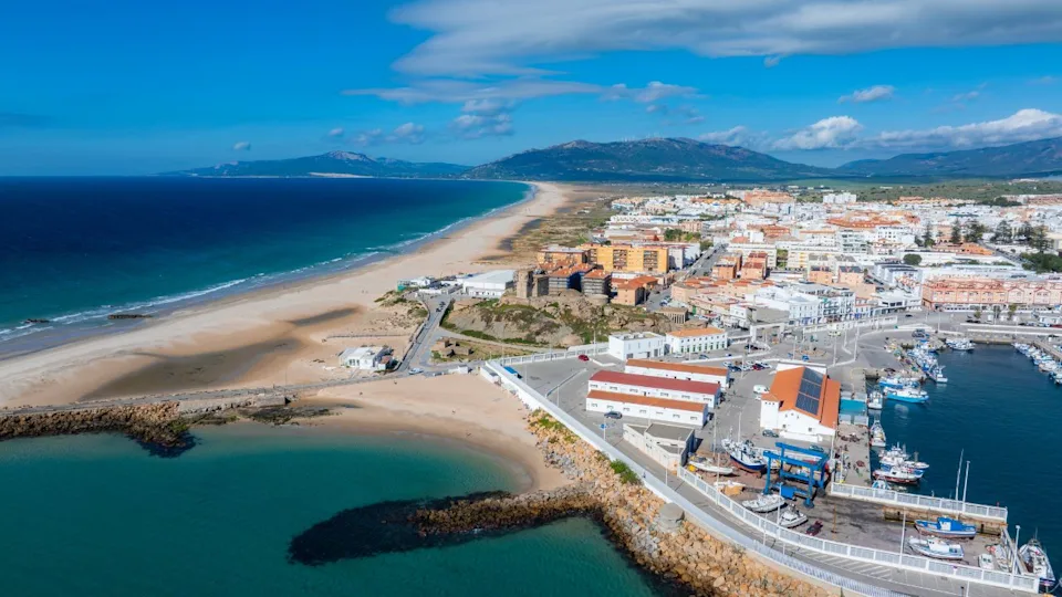 Aerial view of Tarifa, Spain, showing a sandy beach, deep blue Atlantic waters, a marina with boats, town buildings, rolling hills, and a clear sky.