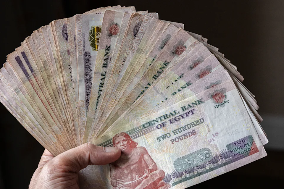 This close-up image shows a hand holding a fanned-out stack of Egyptian two hundred pound banknotes, issued by the Central Bank of Egypt. Prominently displayed on the front of each note is an image of the Seated Scribe, an iconic figure from ancient Egyptian art, symbolizing knowledge and history. The currency features intricate Arabic calligraphy and detailed patterns, reflecting Egypt's rich cultural and artistic heritage. The photo effectively captures a sense of economic value and local identity, making it a powerful visual for topics related to finance, inflation, or the Egyptian economy.