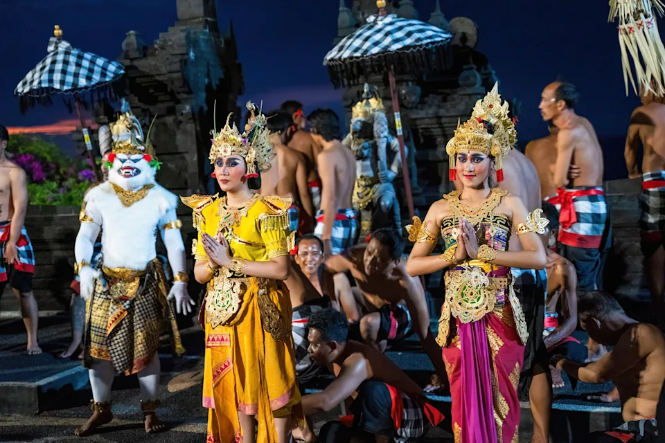 Bali, Indonesia - November 23, 2023: Balinese performers in golden costumes pose gracefully during a Kecak dance, as Hanuman and chanting men create a backdrop at ancient temple under the night sky.