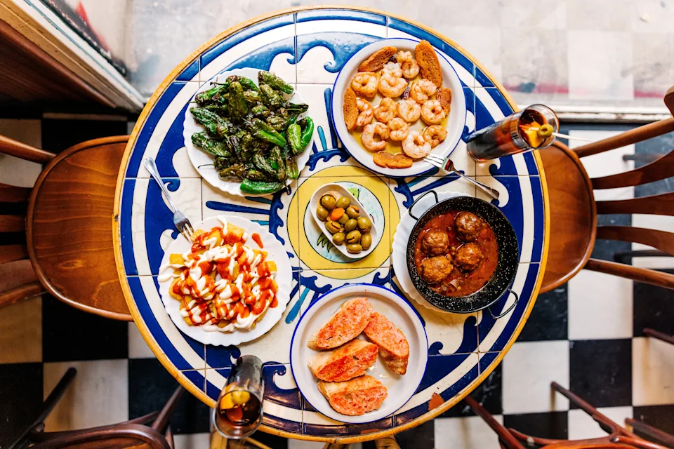 An overhead view of a table with various Spanish tapas, including grilled peppers, shrimp, olives, meatballs, and toasted bread, set for dining