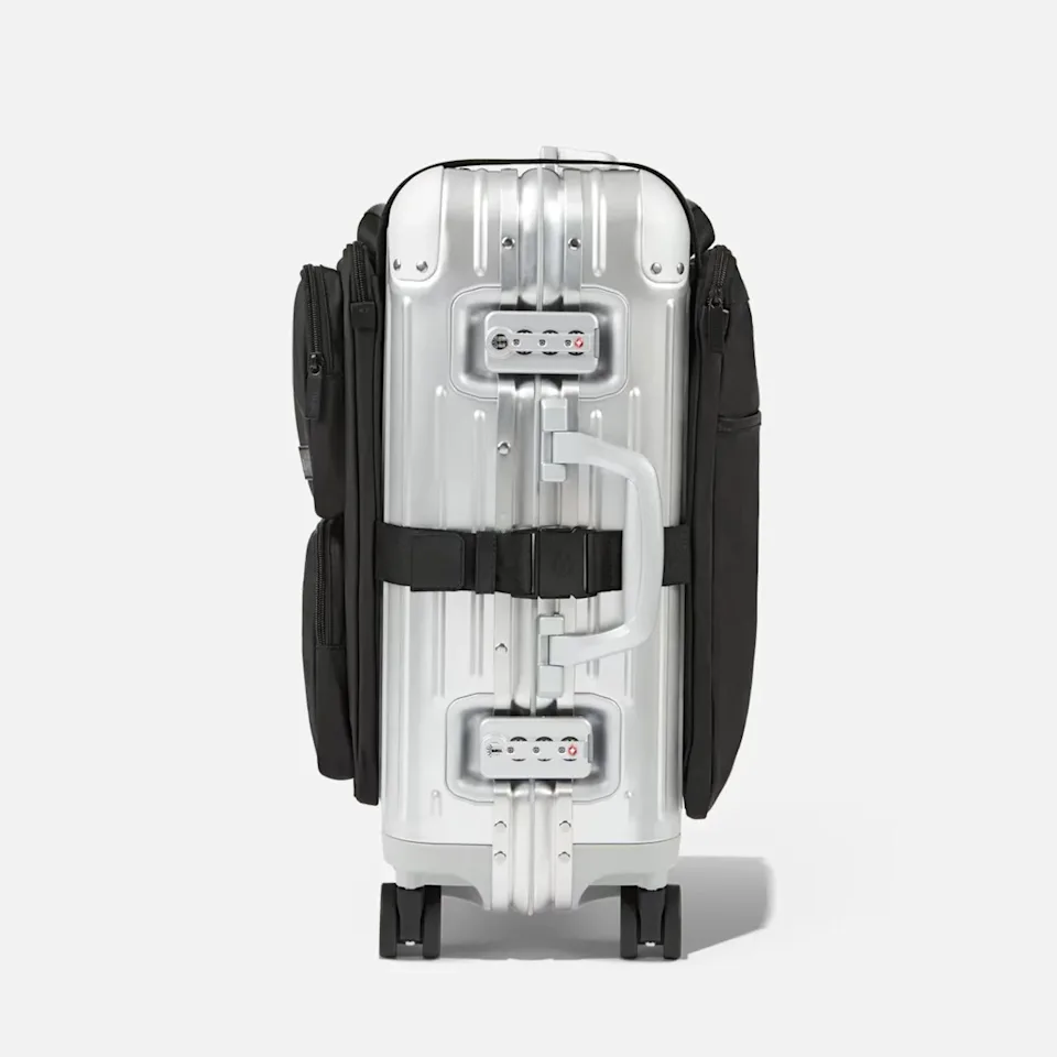 Silver hard-shell suitcase with black fabric side pockets and a black strap around the middle, shown from the side.