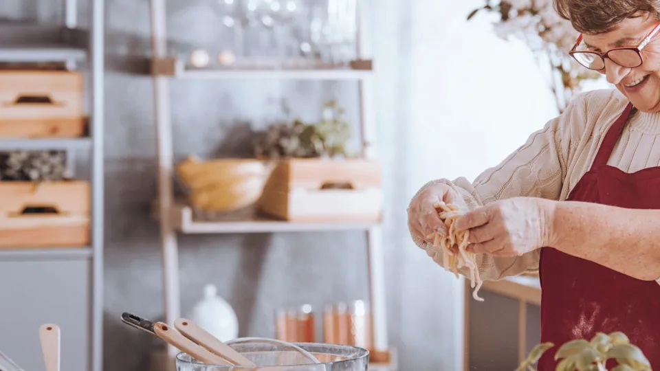 Cheerful senior woman wearing apron cooking in the kitchen enjoying preparing delicious homemade pasta