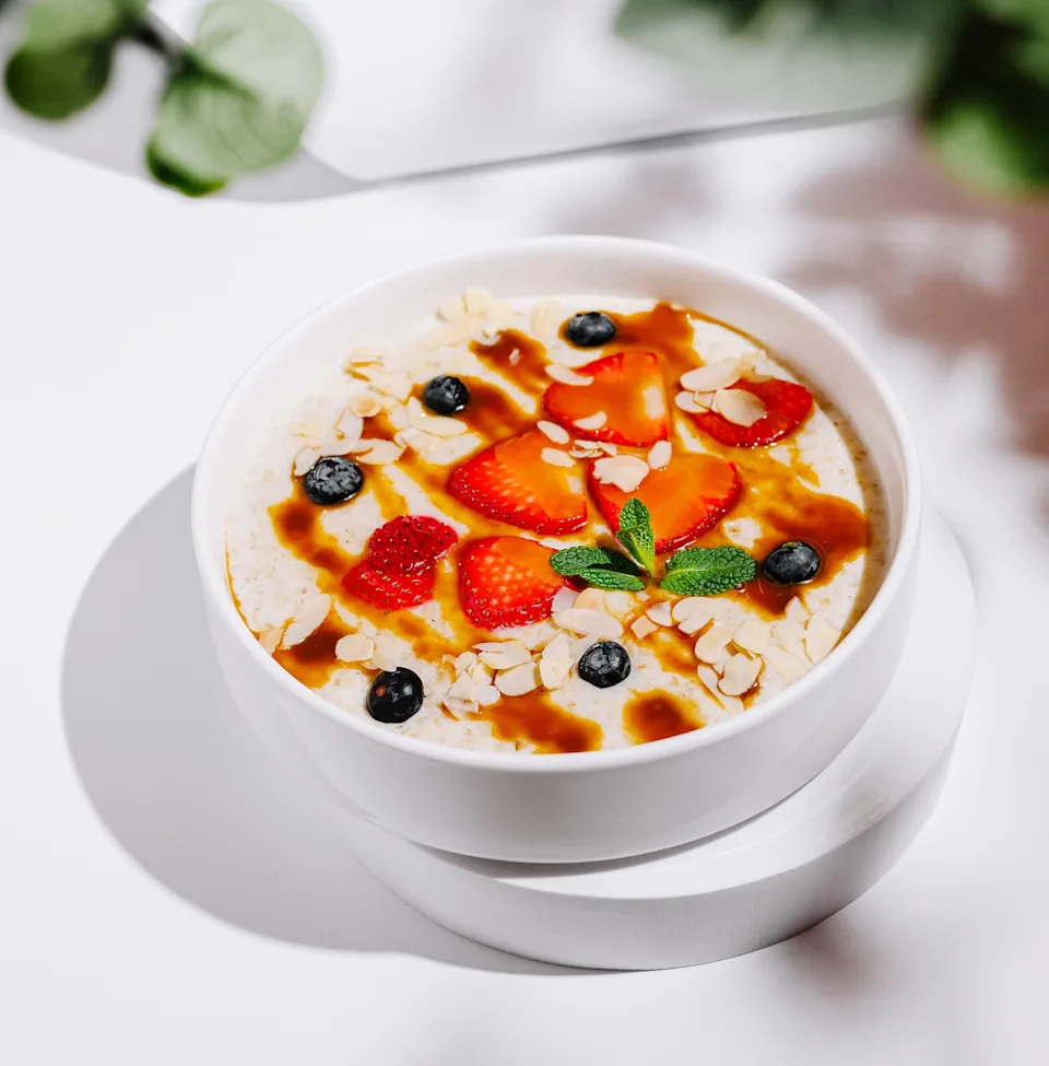 Bowl of oatmeal topped with sliced strawberries, blueberries, almond slivers, mint, and drizzle, set on a white background with subtle greenery