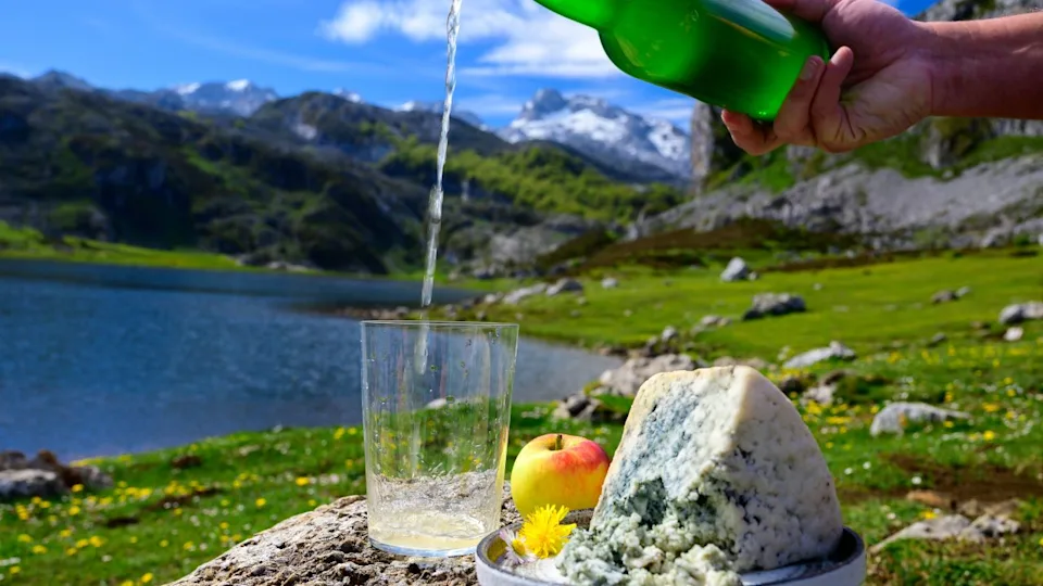 Pouring in glass of natural Asturian cider made from fermented apples, Asturian cabrales cow blue cheese with view on Covadonga lakes and tops of Picos de Europa mountains, Spain