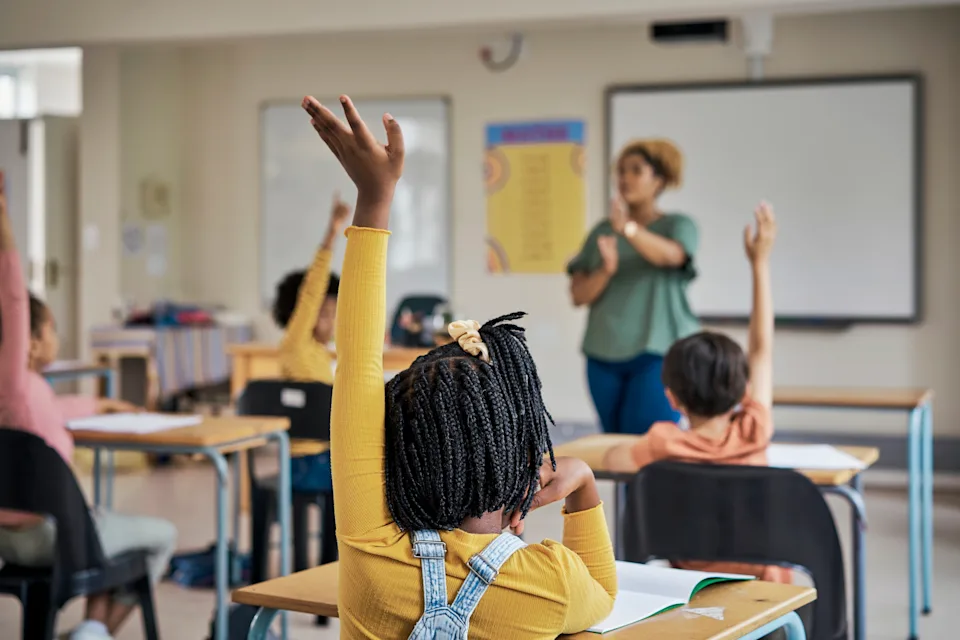 Students in a classroom raise their hands, engaging with the teacher standing at the front