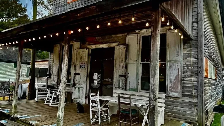 A weathered wooden building, wooden table and chairs on the porch, shutters on the window