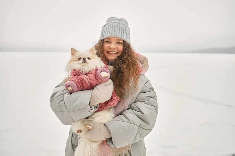 Portrait of Caucasian young adult woman smiling while holding small fluffy dog dressed in warm clothing outdoors on snowy frozen lake during winter season